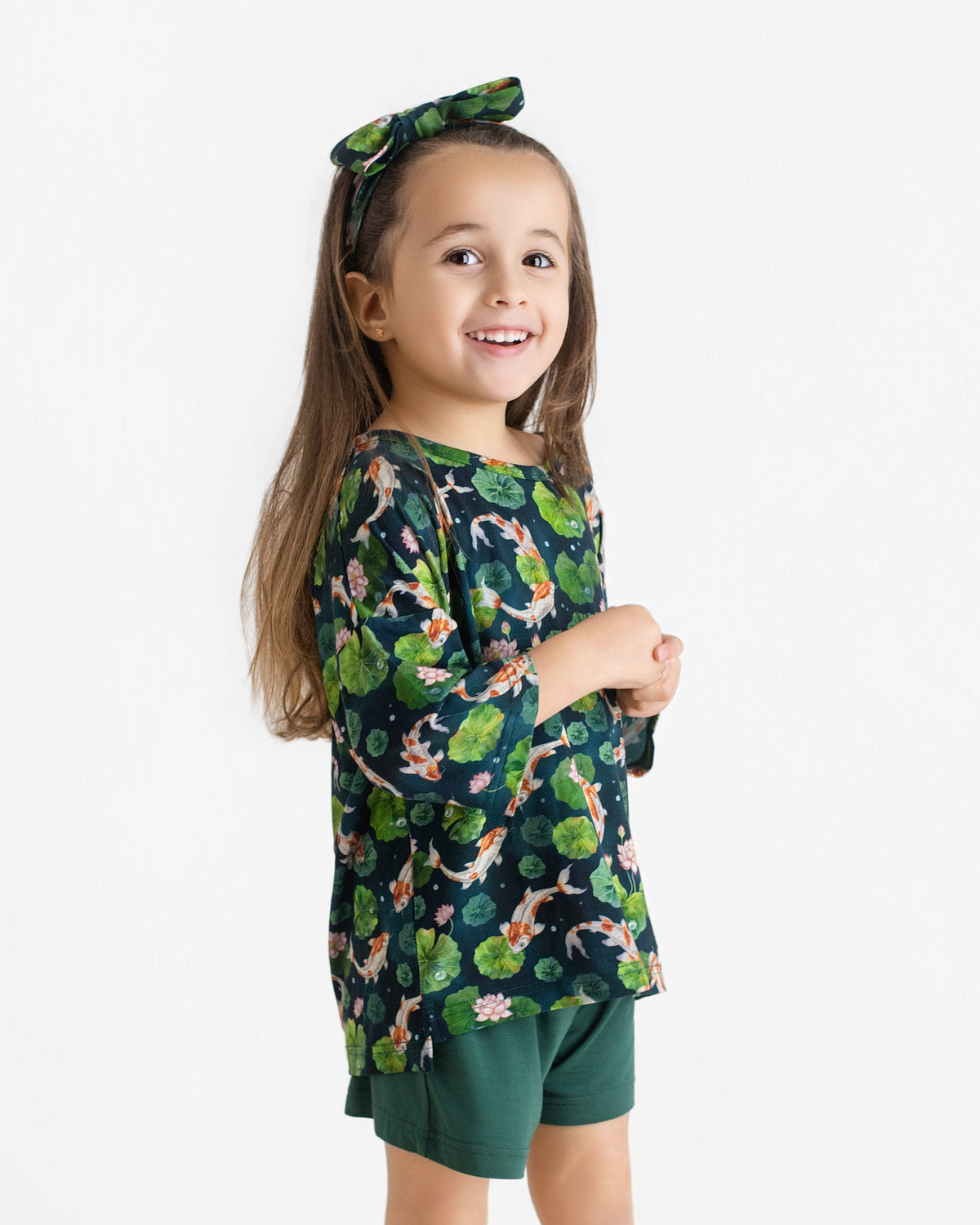 A young girl smiles in the Jammie Session Bundle of Koi Comfy Cloud Short Set—dark green floral top with matching headband and green shorts—standing against a plain white background.