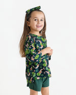 A young girl smiles in the Jammie Session Bundle of Koi Comfy Cloud Short Set—dark green floral top with matching headband and green shorts—standing against a plain white background.