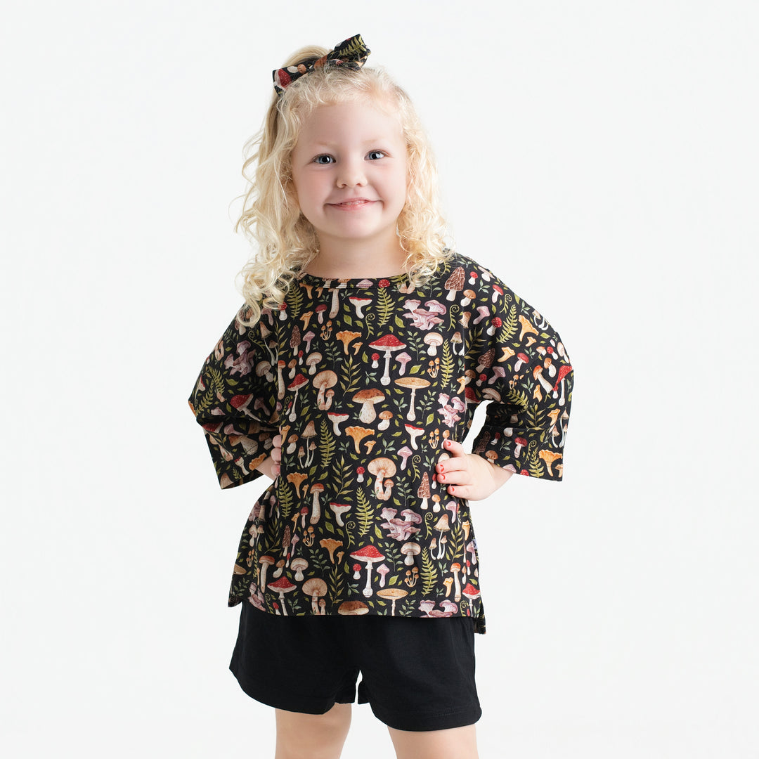 A young girl with blonde curly hair wears the Mystic Mushrooms Comfy Cloud Short Set by Jammie Session, featuring a mushroom-patterned black top, matching shorts, and hair bow, standing against a plain white background.
