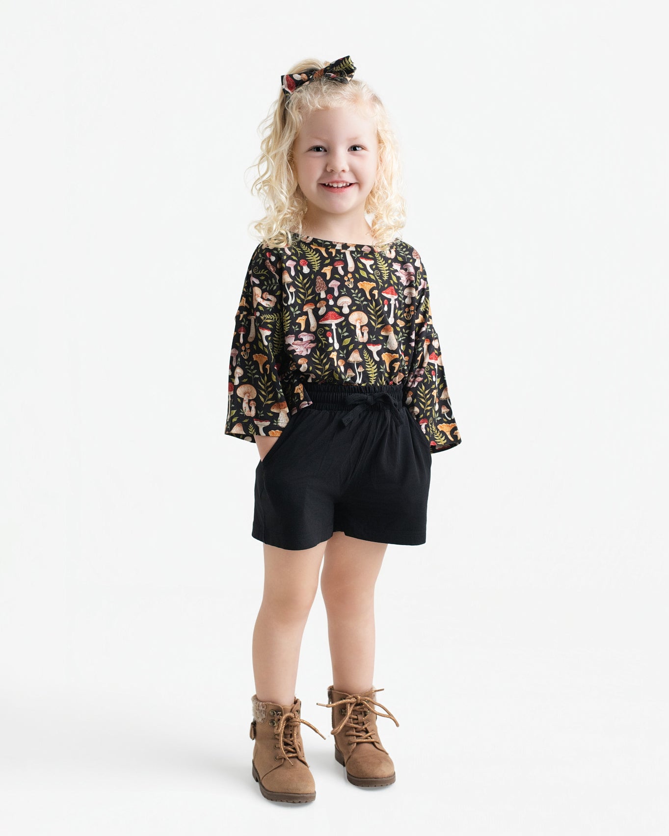 A young child smiles in the Jammie Session Mystic Mushrooms Comfy Cloud Short Set, featuring a patterned top and black shorts, with brown boots and a matching hair bow, posed against a plain white background.