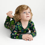 A young child with curly blond hair and blue eyes lies on their stomach in Jammie Session's Bundle of Koi Long Sleeve PJ’s, featuring a floral and animal print, against a plain white background.