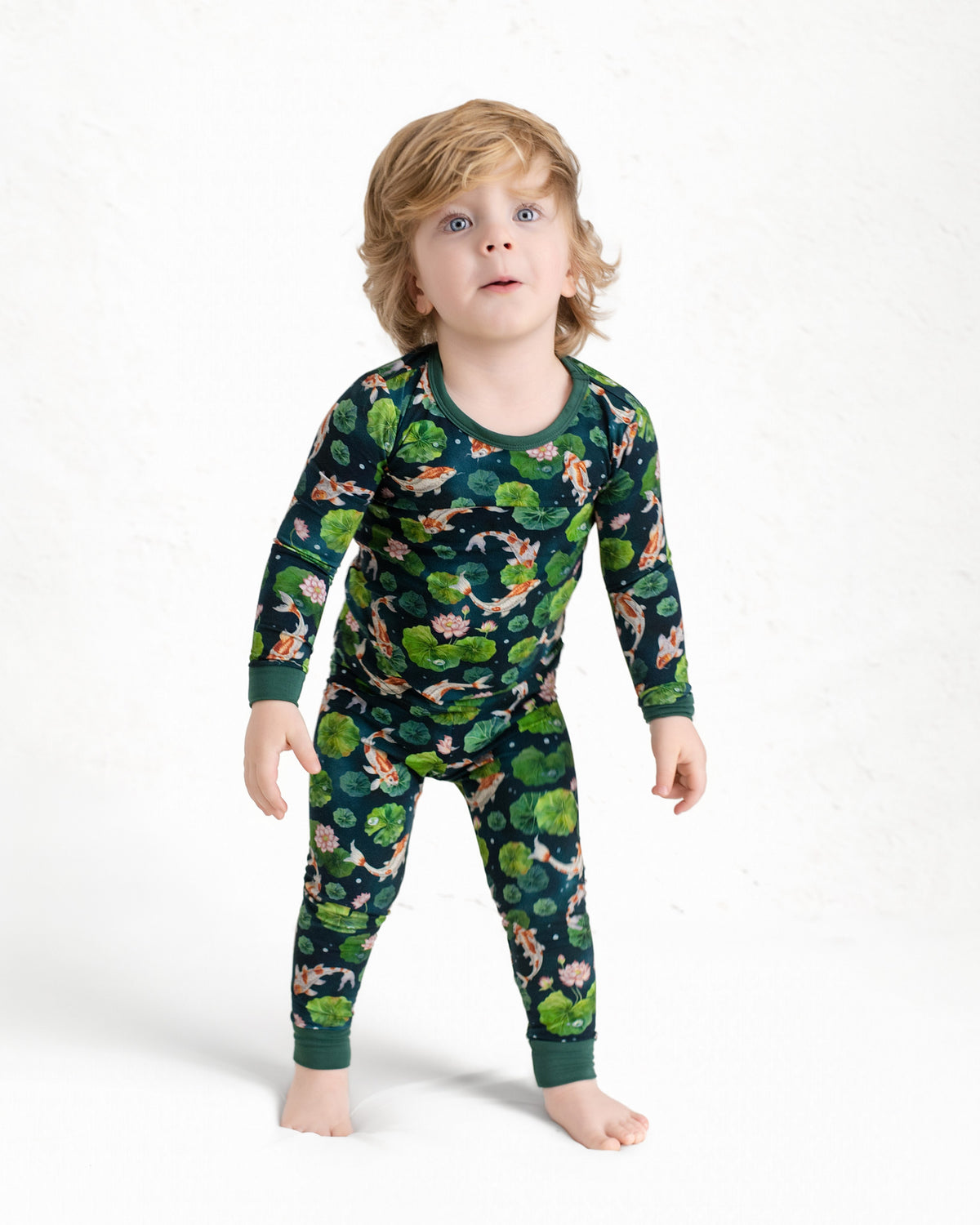 A young child with light brown hair stands barefoot against a white background wearing Jammie Session’s Bundle of Koi Long Sleeve PJ's—hypoallergenic kids pajamas in dark green with a nature and animal print.