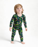 A young child with light brown hair stands barefoot against a white background wearing Jammie Session’s Bundle of Koi Long Sleeve PJ's—hypoallergenic kids pajamas in dark green with a nature and animal print.
