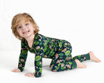 A young blond child, smiling at the camera, crawls on a white background while wearing Jammie Session’s Bundle of Koi Long Sleeve PJ's featuring a dark green color with floral and animal patterns.