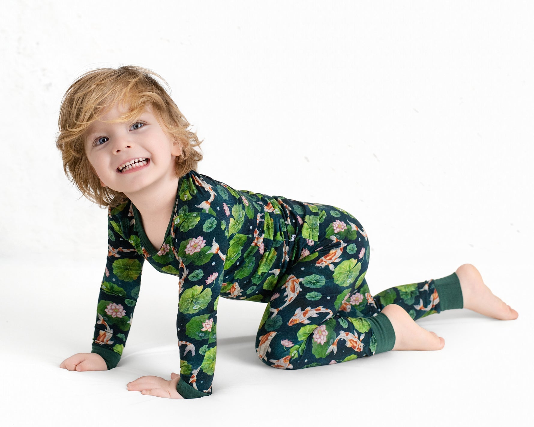 A young blond child, smiling at the camera, crawls on a white background while wearing Jammie Session’s Bundle of Koi Long Sleeve PJ's featuring a dark green color with floral and animal patterns.