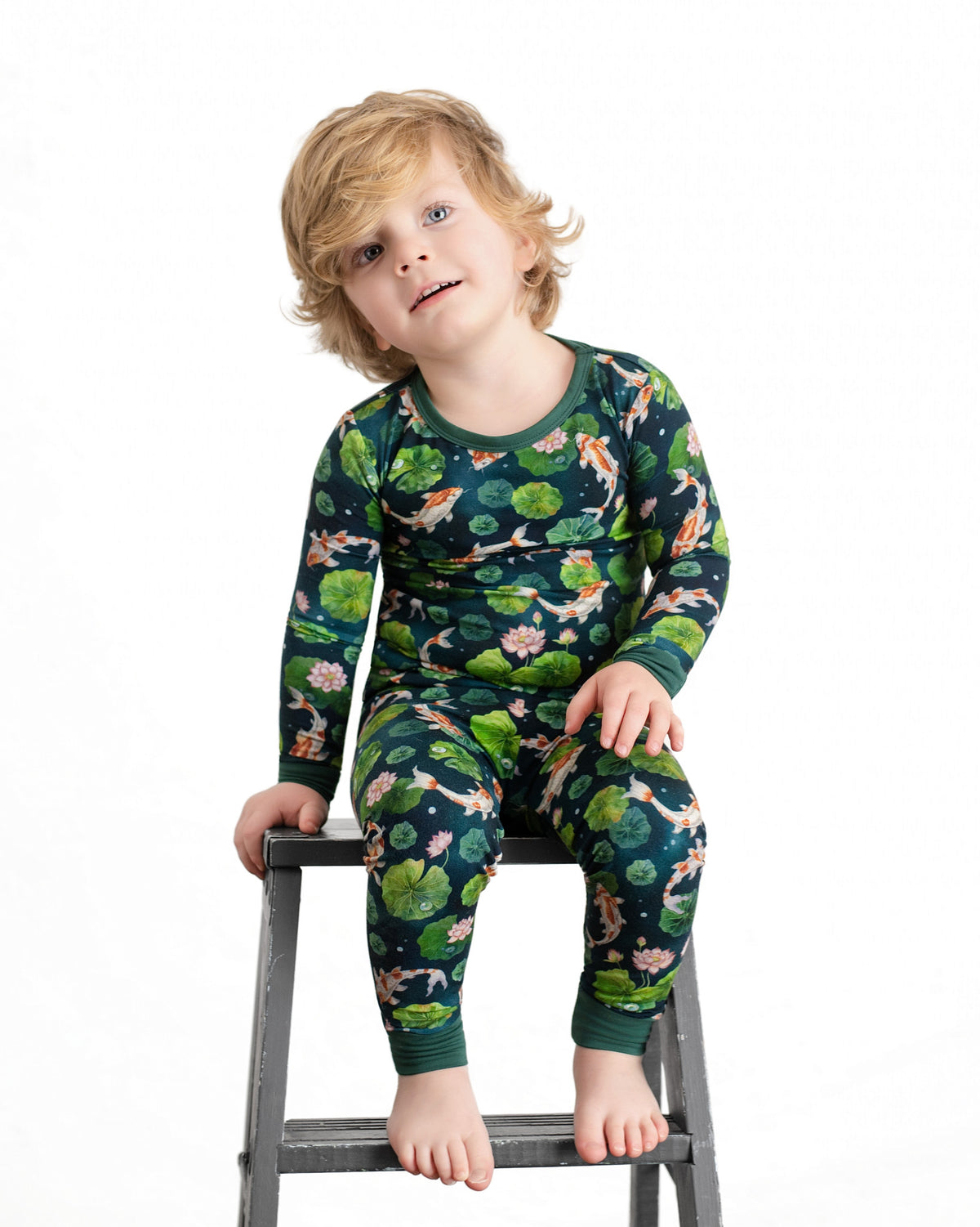 A young blond child sits barefoot on a gray step ladder, wearing matching dark green Bundle of Koi Long Sleeve PJ's by Jammie Session, against a plain white background.