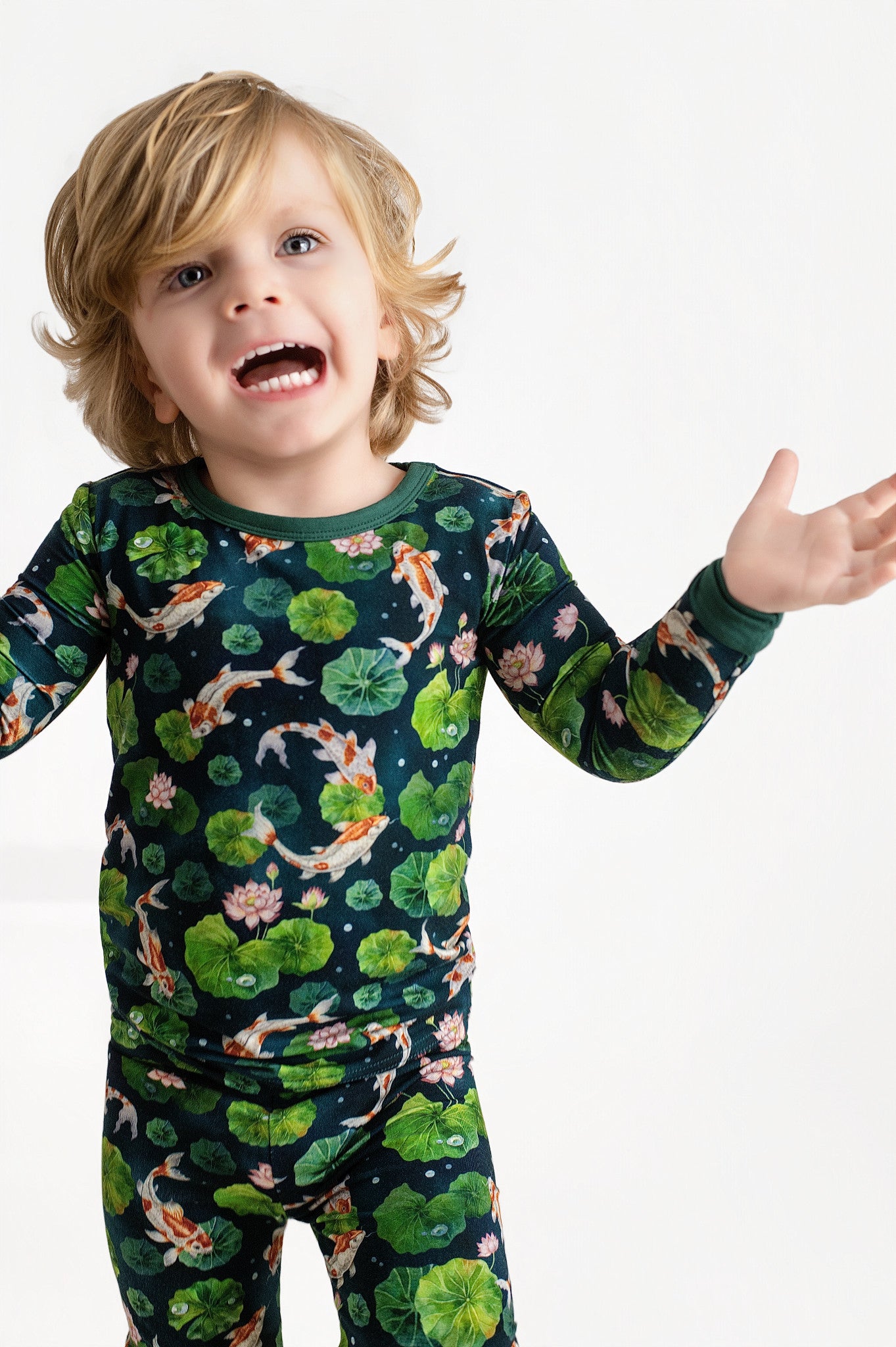 A young child with blonde hair wears Jammie Session’s Bundle of Koi Long Sleeve PJ's, featuring hypoallergenic fabric and a koi fish and lily pad print, standing against a plain white background with arms raised and mouth open.