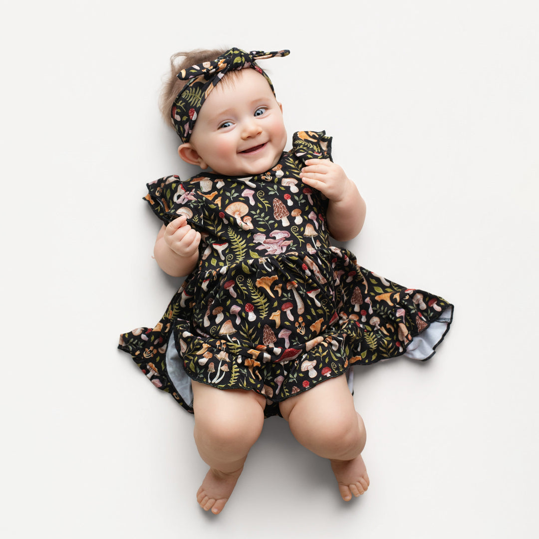 A baby smiles on a white background, wearing the Jammie Session Mystic Mushrooms Bodysuit Twirl Dress and matching headband. This bamboo dress is soft and designed for easy diaper changes, offering everyday comfort.