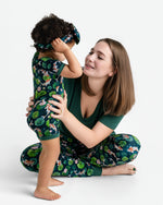 A woman smiles at a toddler in front of her, both wearing Jammie Session matching floral and fish print outfits. The child wears the Bundle of Koi Short Sleeve Romper with a matching headband, against a white background.