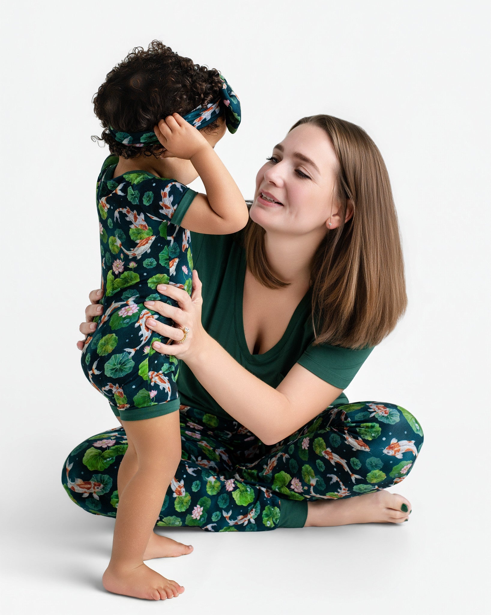 A woman smiles at a toddler in front of her, both wearing Jammie Session matching floral and fish print outfits. The child wears the Bundle of Koi Short Sleeve Romper with a matching headband, against a white background.