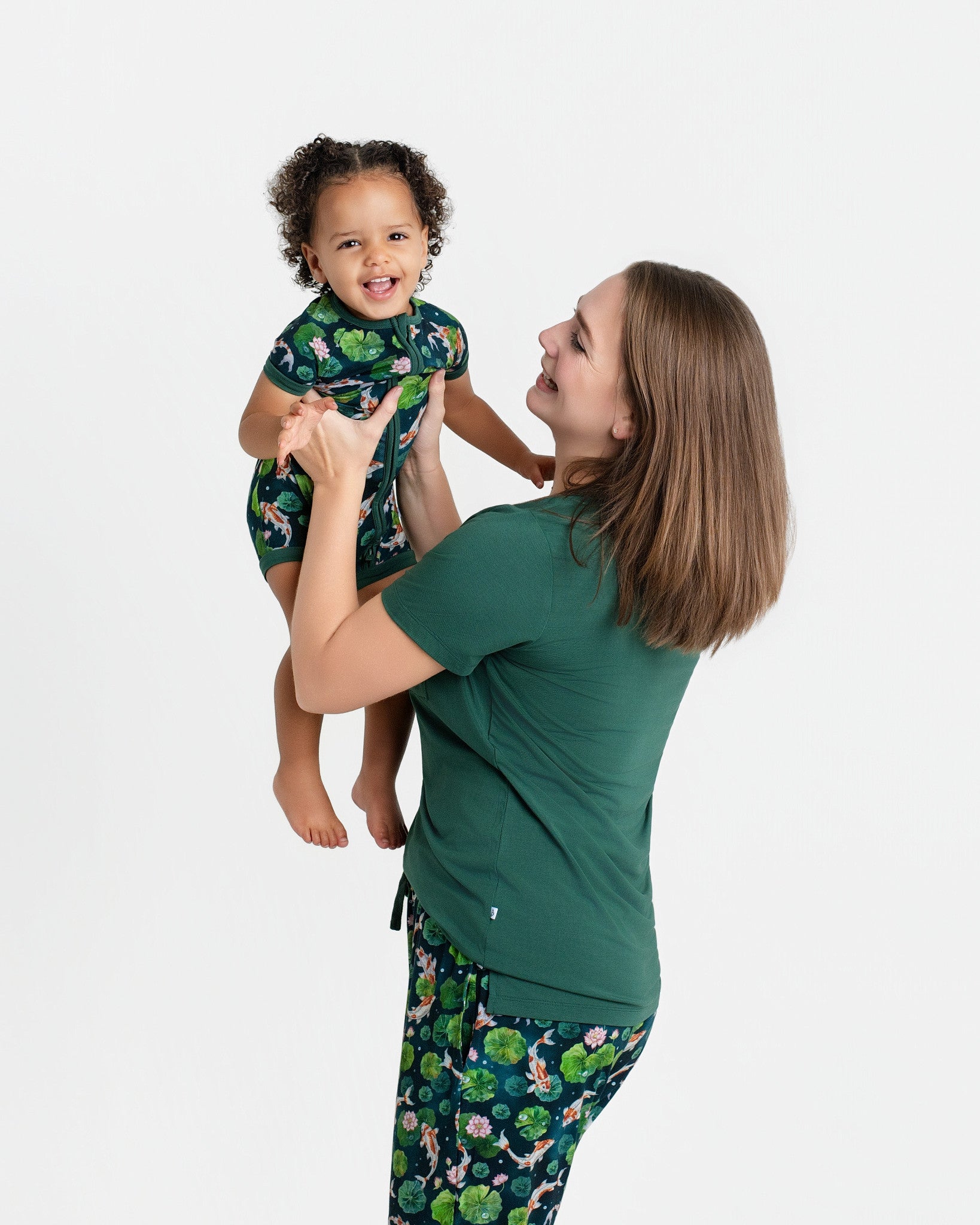 A woman in a green Jammie Session Bundle of Koi Women's Solid Short Sleeve T-shirt lifts a smiling toddler in matching pajamas against a plain white background.