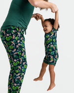 An adult holds a smiling child by the arms; both wear matching Jammie Session "Bundle of Koi" short sleeve rompers, featuring dark green bamboo fabric with colorful floral and fish patterns, set against a white background.
