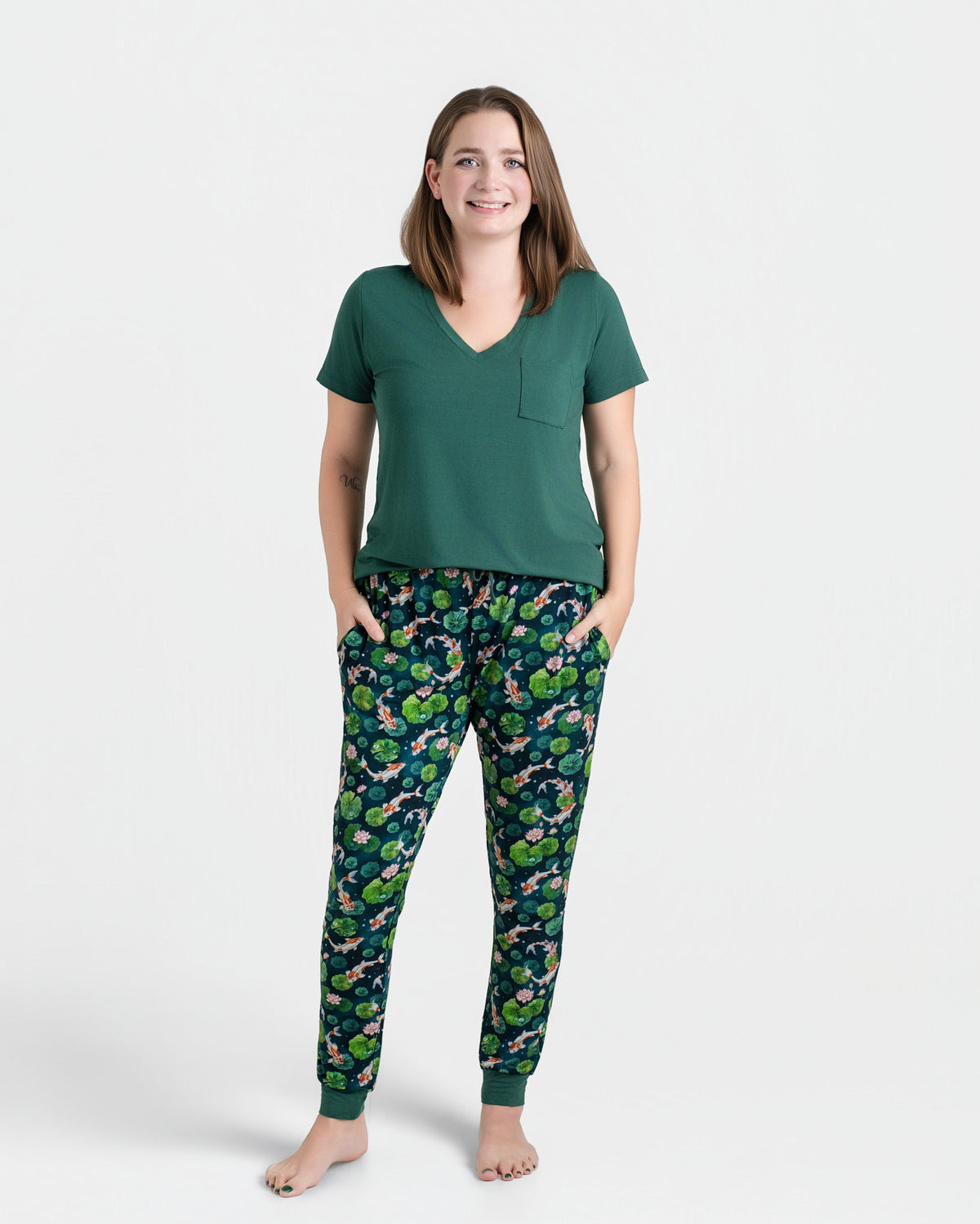A woman stands barefoot in a Jammie Session Bundle of Koi Women's Solid Short Sleeve T-shirt and patterned pajama pants, hands in pockets. The moisture-wicking fabric keeps her comfortable all day against a plain white background.