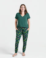 A woman stands barefoot in a Jammie Session Bundle of Koi Women's Solid Short Sleeve T-shirt and patterned pajama pants, hands in pockets. The moisture-wicking fabric keeps her comfortable all day against a plain white background.