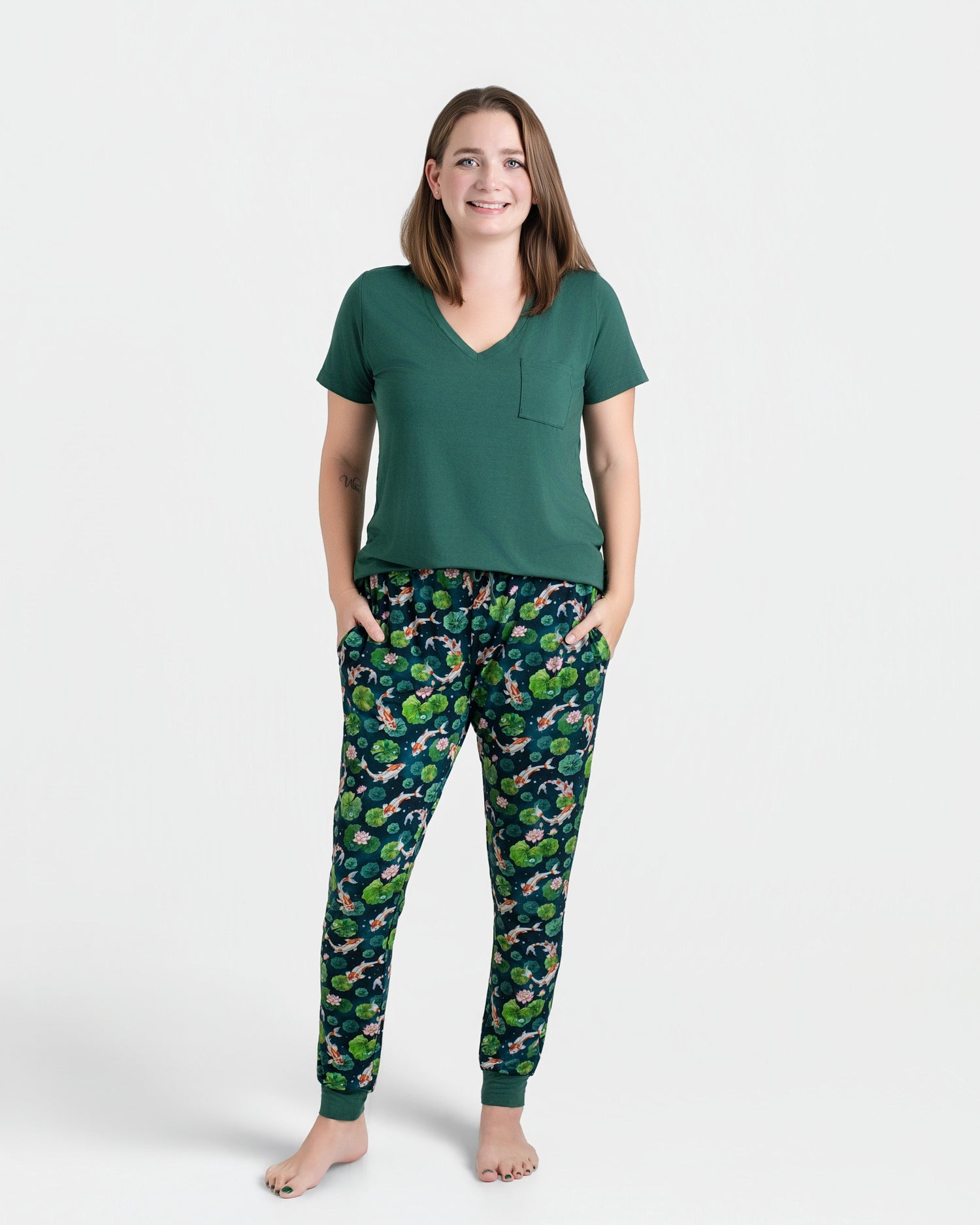 A woman stands barefoot in a Jammie Session Bundle of Koi Women's Solid Short Sleeve T-shirt and patterned pajama pants, hands in pockets. The moisture-wicking fabric keeps her comfortable all day against a plain white background.