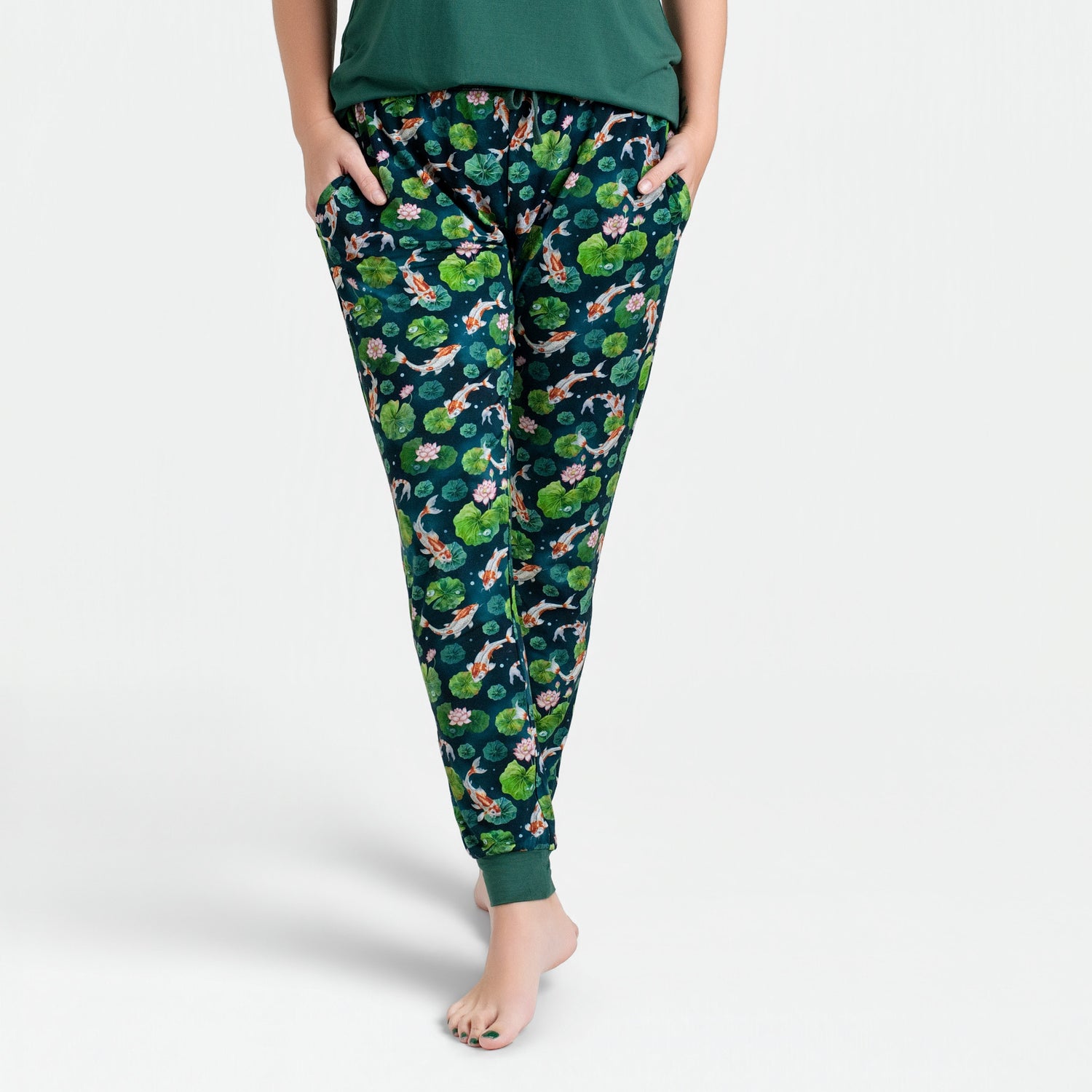 A woman wears Jammie Session's Bundle of Koi Women's Jogger Pants—dark green with a vibrant floral and animal print—hands in pockets, standing barefoot against a white background.