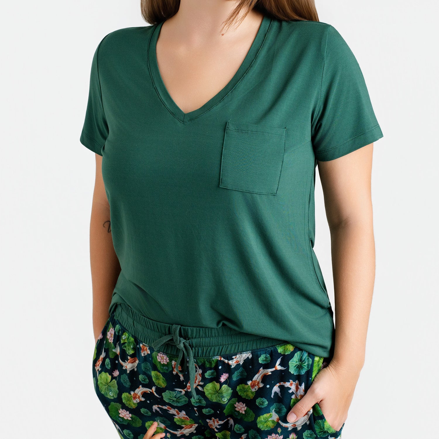 A woman wears the Jammie Session Bundle of Koi Women's Solid Short Sleeve T-shirt with V-neck and chest pocket, paired with drawstring animal-and-leaf print pants. The tee is breathable and moisture wicking for all-day comfort.