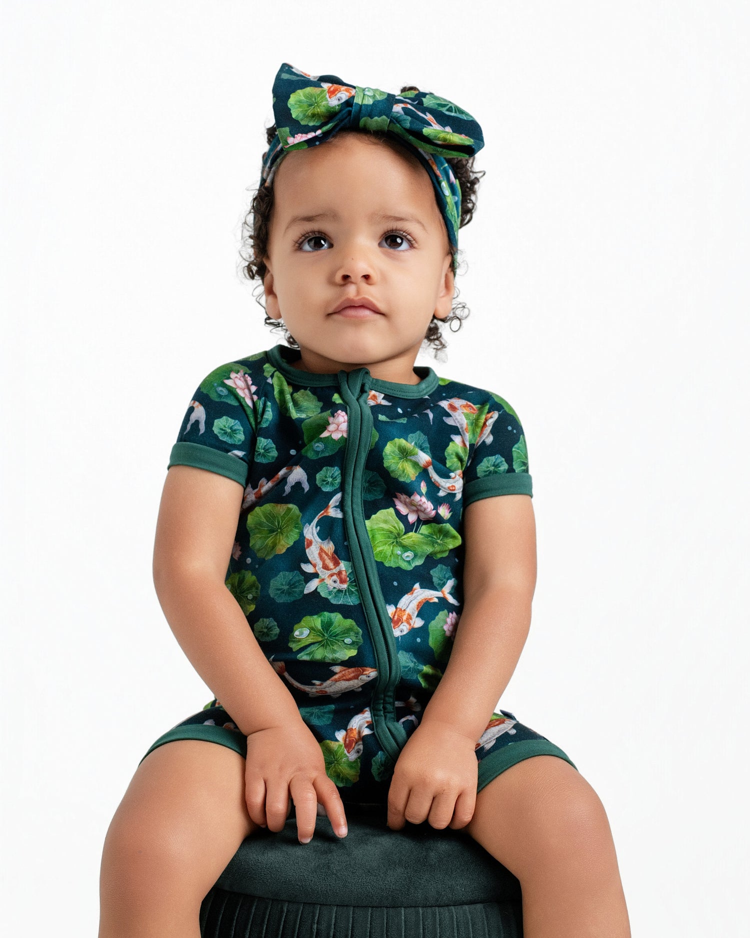 A toddler models the Jammie Session Bundle of Koi Short Sleeve Romper with a matching headband, featuring koi fish and lotus flowers, while seated on a dark green stool against a white background.