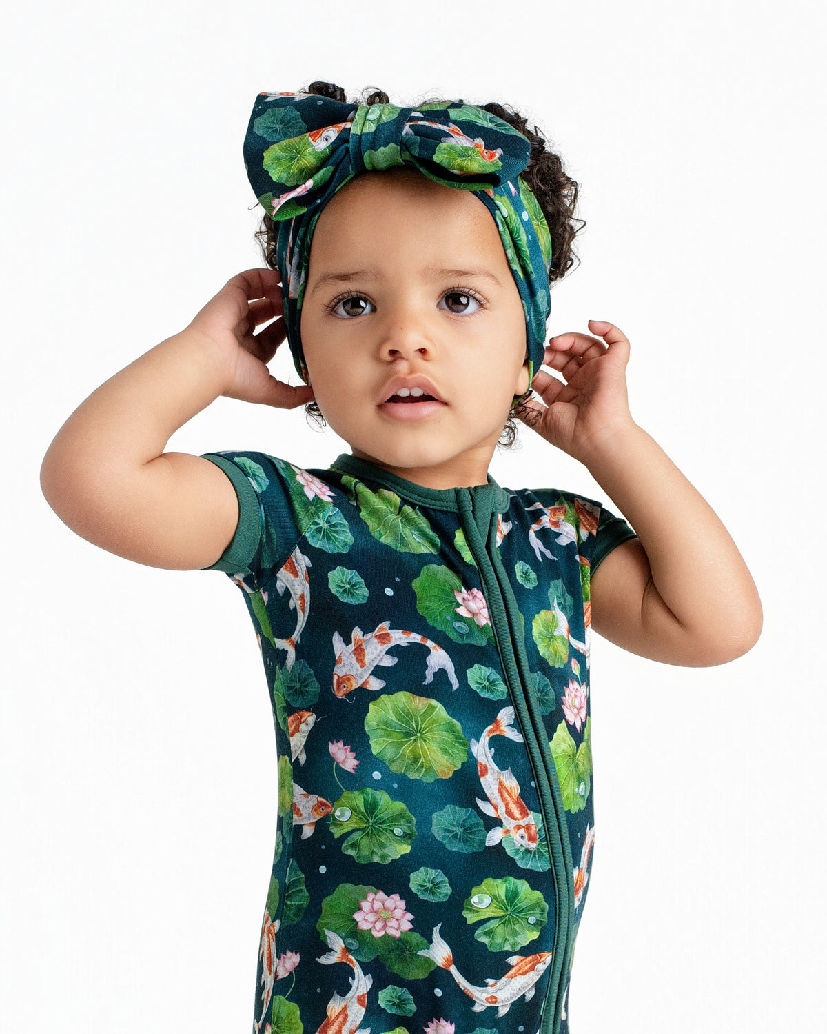 A toddler in the Jammie Session Bundle of Koi Short Sleeve Romper and matching green headband with koi fish and lily pad print stands against a plain white background.