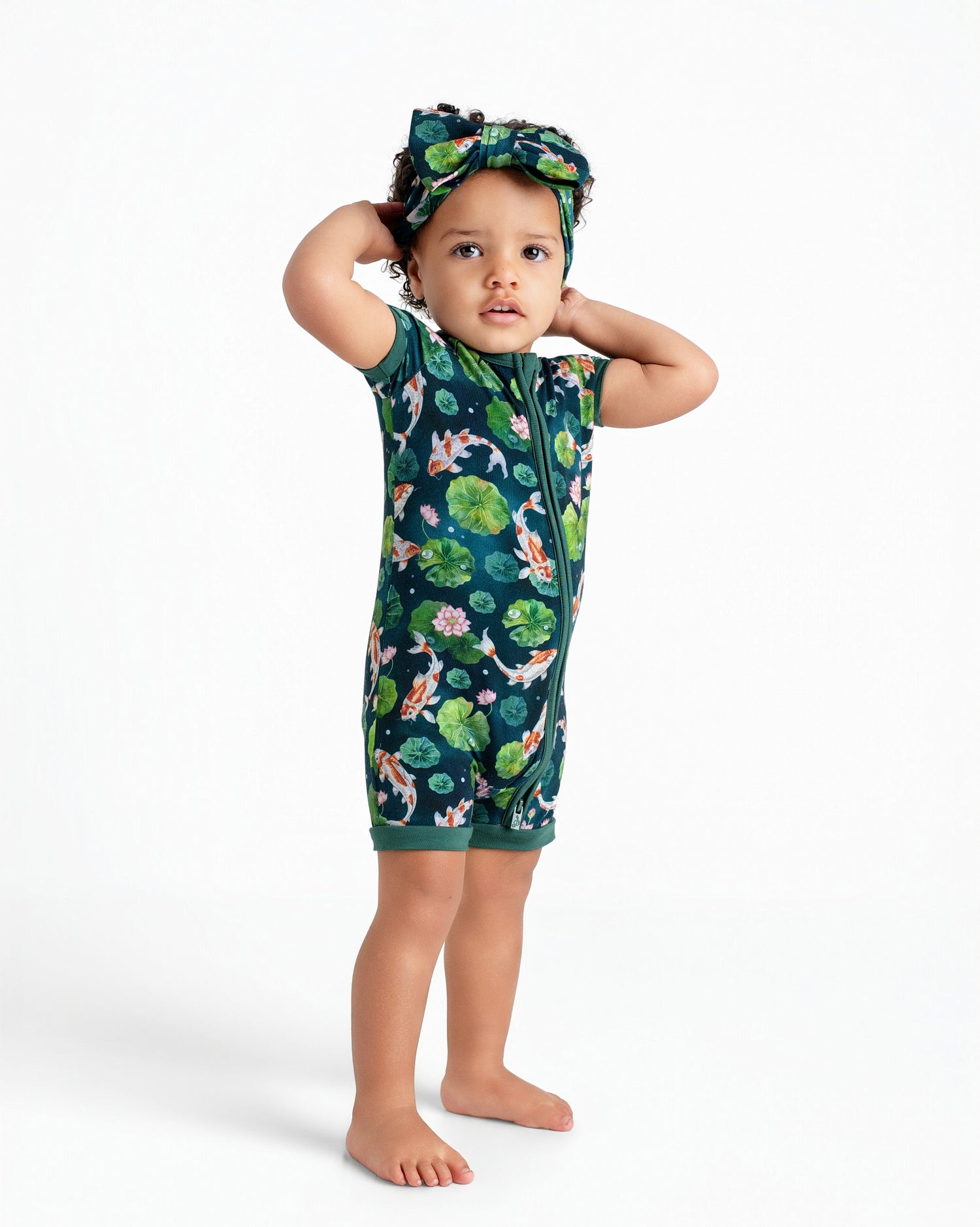A toddler stands barefoot with hands behind head, dressed in the Jammie Session Bundle of Koi Short Sleeve Romper and matching headband, against a plain white background.