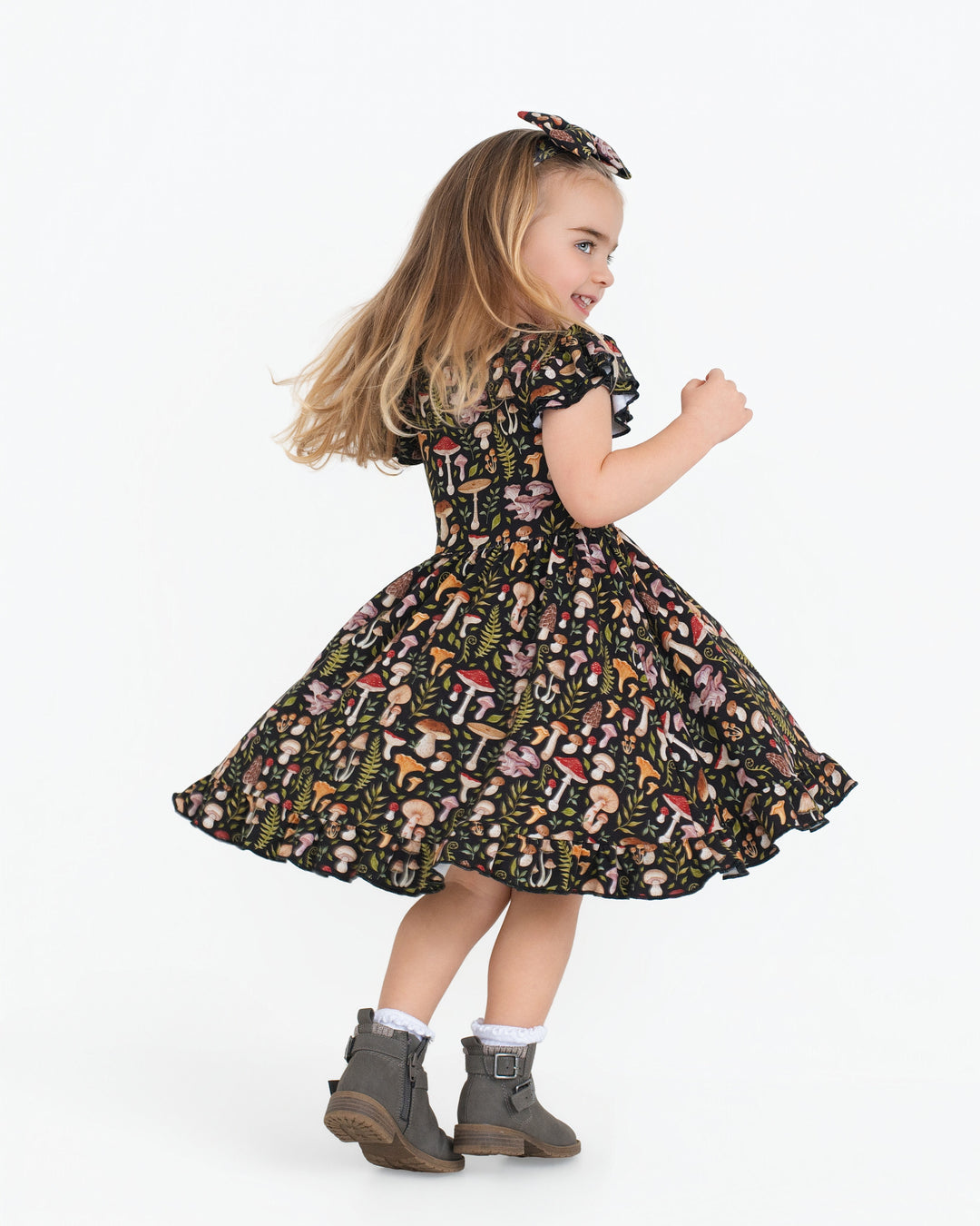 A young girl wears the Jammie Session Mystic Mushrooms Short Sleeve Big Kid Twirl Dress, paired with gray boots and a headband, standing sideways against a plain white background.
