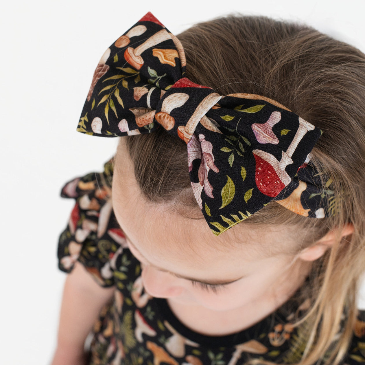 A young child with light brown hair wears the Jammie Session Mystic Mushrooms Bow, which features a colorful mushroom pattern that matches the design on her dress sleeves.