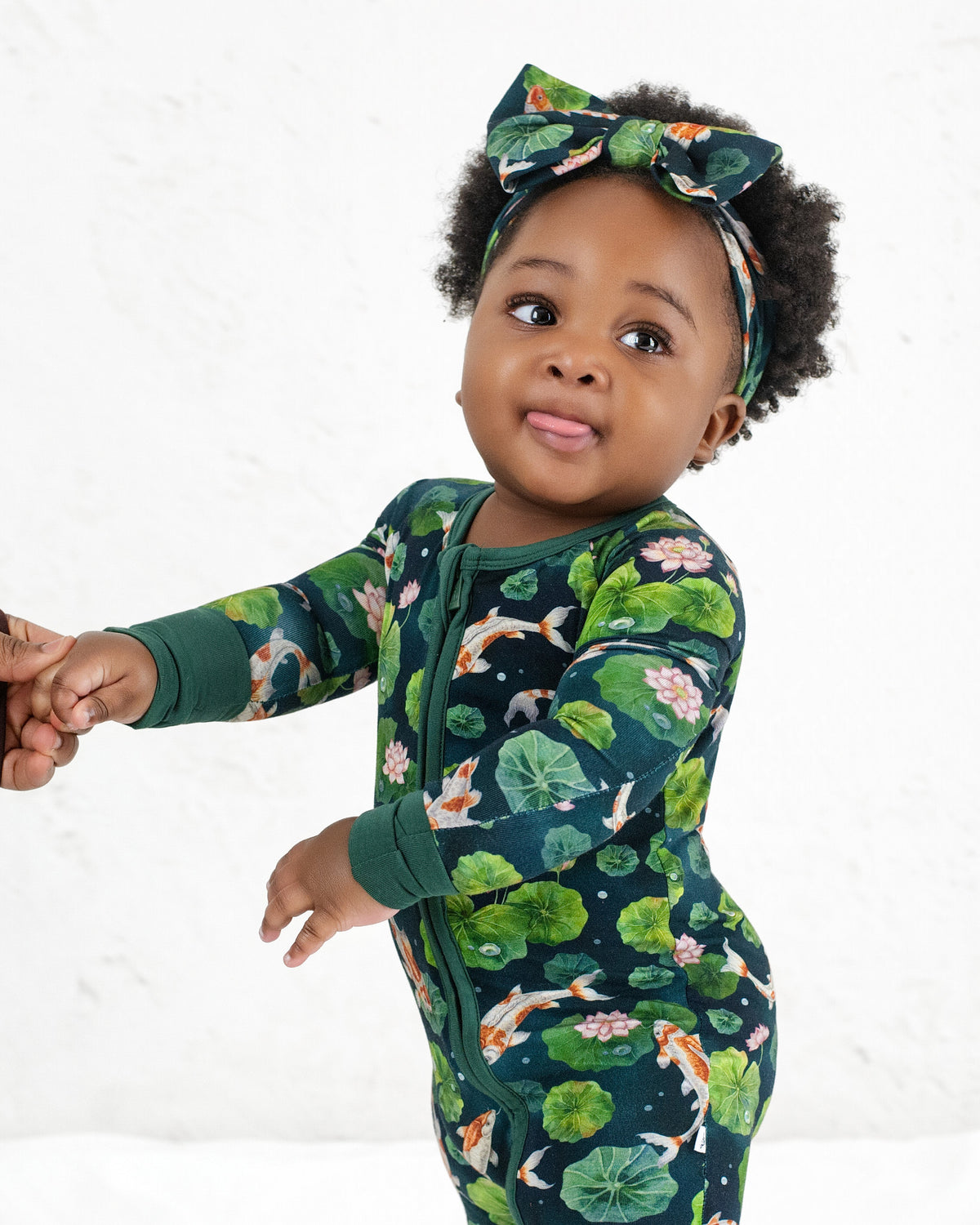 A toddler wears the Jammie Session Bundle of Koi Romper with a matching headband, holds an adult’s hand, and looks ahead with their tongue slightly out.