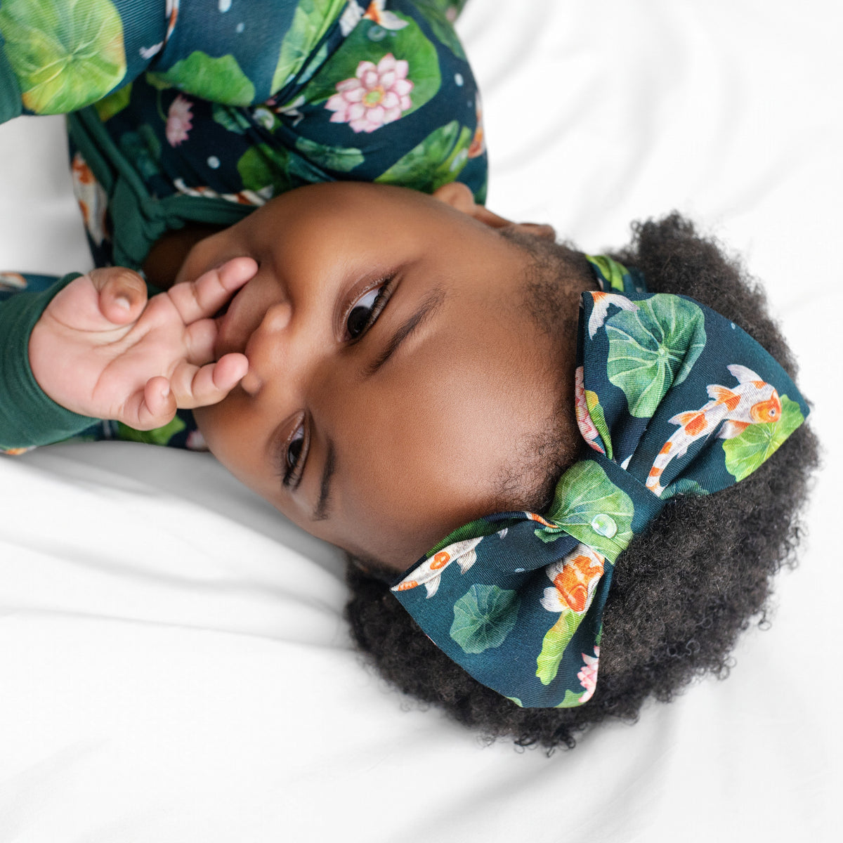 A baby wears the Jammie Session Bundle of Koi Bow, a green floral-patterned outfit with a matching bow headband, lying on a white surface and sucking their thumb while gazing to the side.