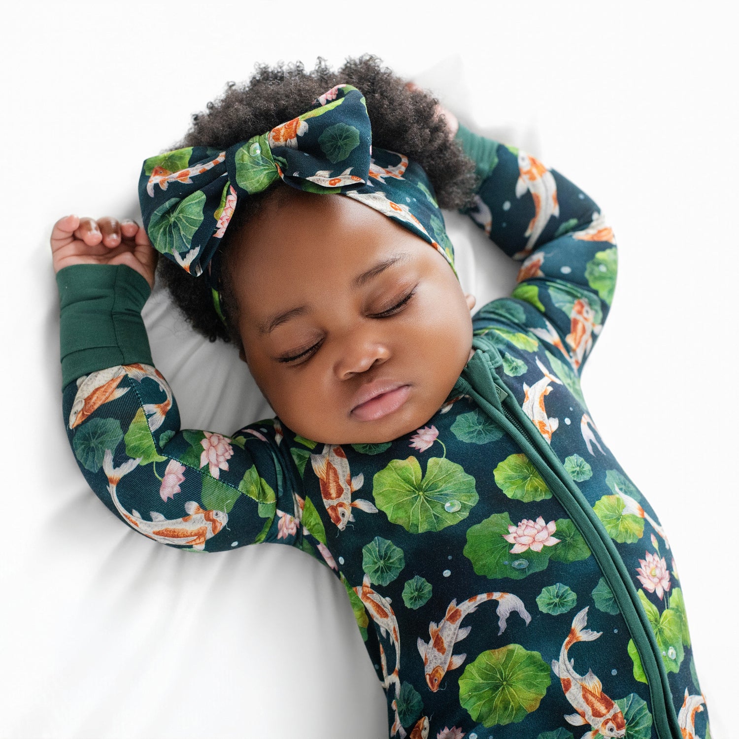 A baby in the Jammie Session Bundle of Koi Romper—a long sleeve bamboo romper with a koi fish and lily pad pattern—wears a matching headband and sleeps on a white surface with arms raised.
