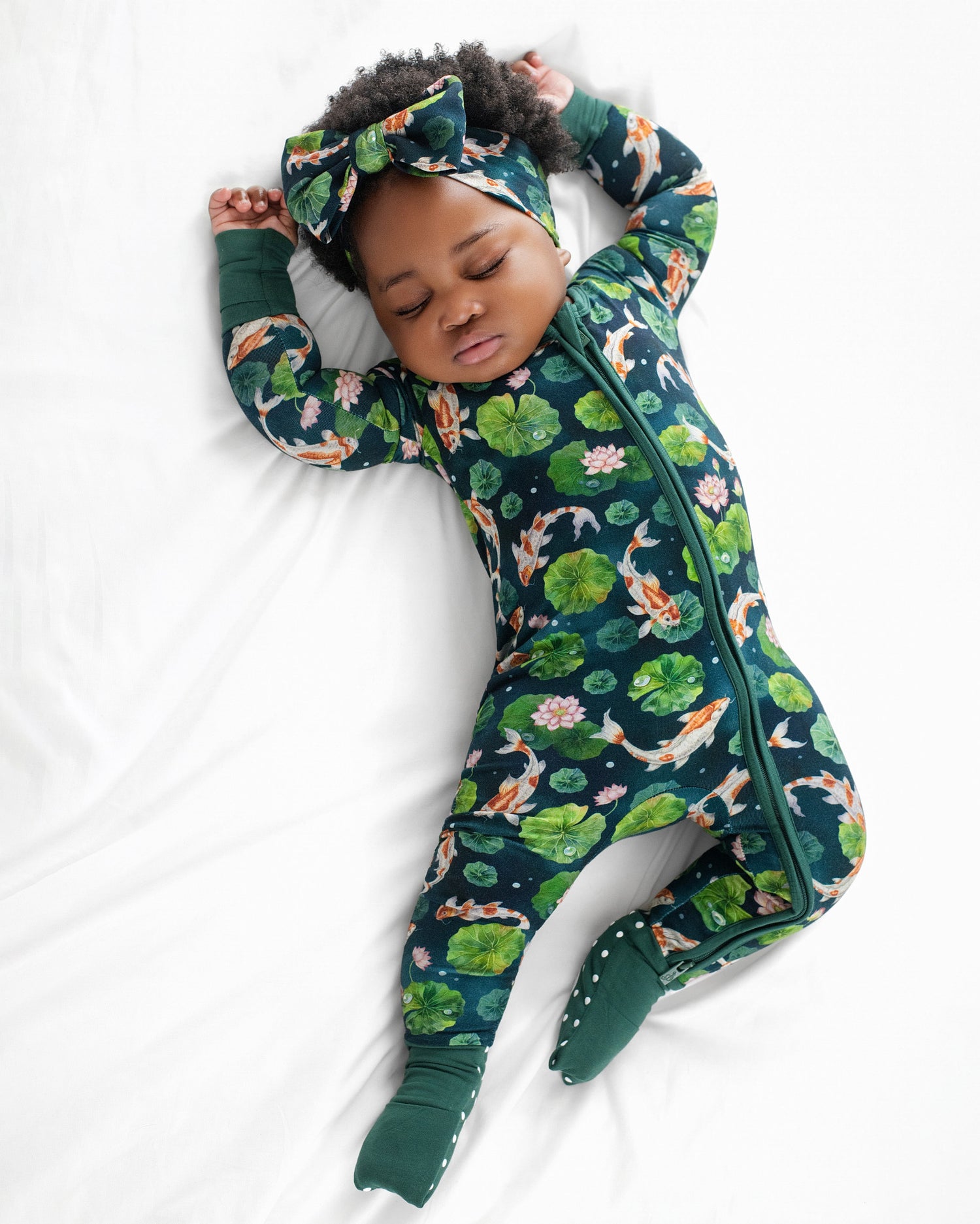 A baby wears the Jammie Session Bundle of Koi Romper—a dark green floral and koi fish patterned convertible footed romper with a matching headband—while sleeping on a white surface.