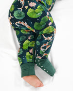 A baby in the Jammie Session Bundle of Koi Romper, featuring a dark green long sleeve bamboo fabric and koi fish with lotus pond pattern, lies on a white surface.