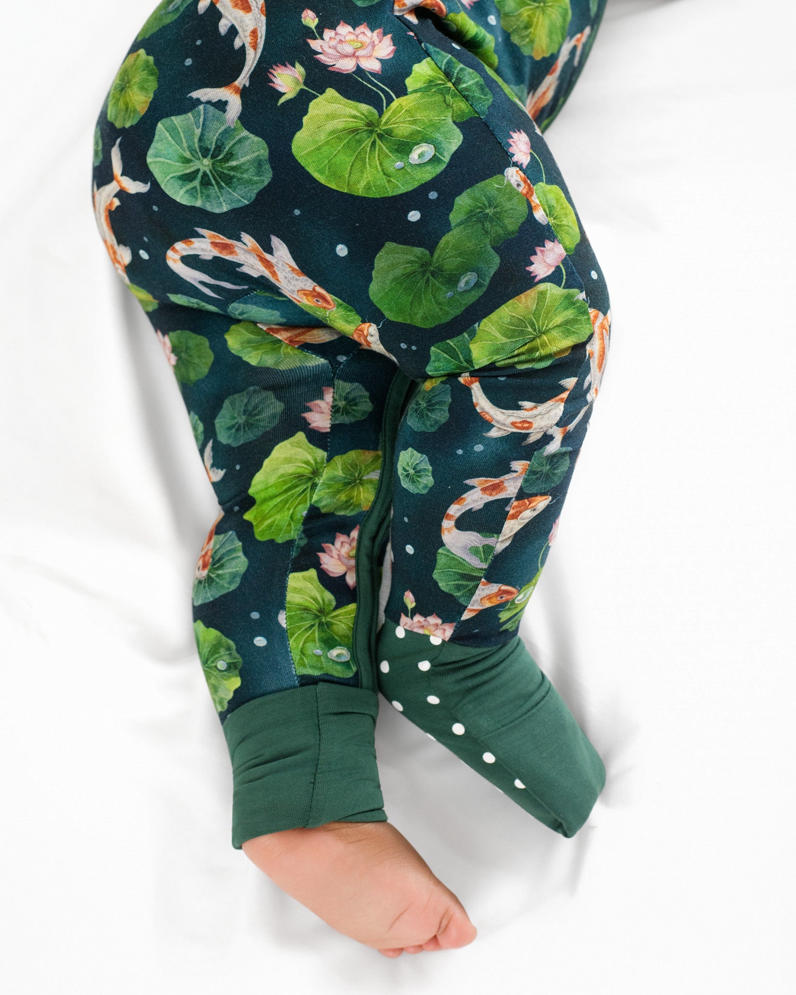 A baby in the Jammie Session Bundle of Koi Romper, featuring a dark green long sleeve bamboo fabric and koi fish with lotus pond pattern, lies on a white surface.