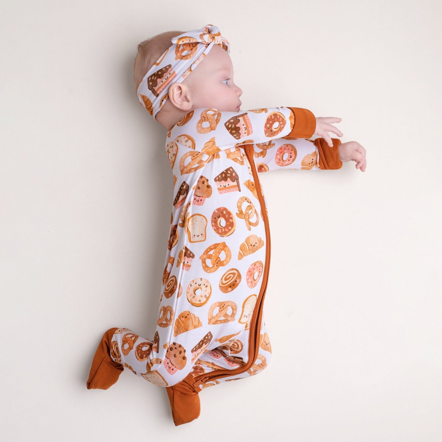 A baby wears the Jammie Session Doughzing Off Romper—a long-sleeve bamboo outfit with matching headband, featuring baked goods prints—lying on a light surface, sideways with arms extended forward.