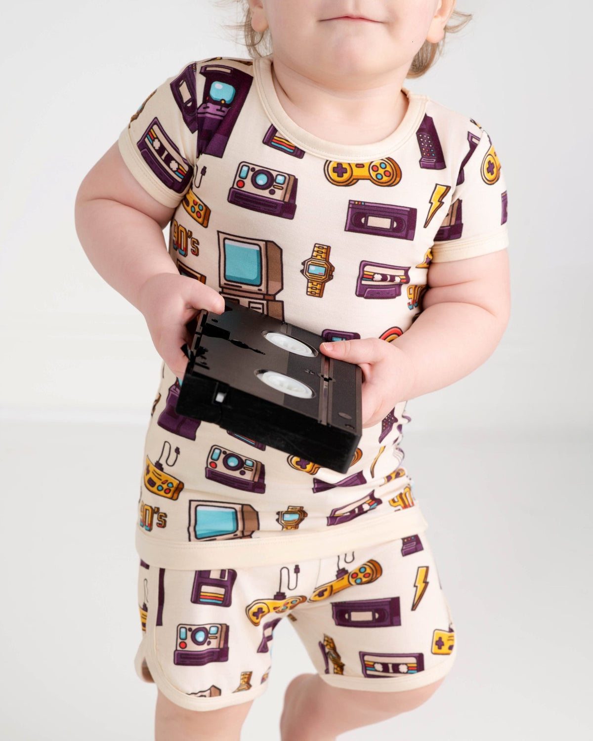 A toddler in the Jammie Session 90's Retro Short Pajama Set—breathable bamboo pajamas with fun TV, watch, game console, and cassette tape prints—holds a VHS tape. Perfect for sensitive skin.