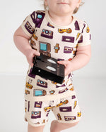 A toddler in the Jammie Session 90's Retro Short Pajama Set—breathable bamboo pajamas with fun TV, watch, game console, and cassette tape prints—holds a VHS tape. Perfect for sensitive skin.