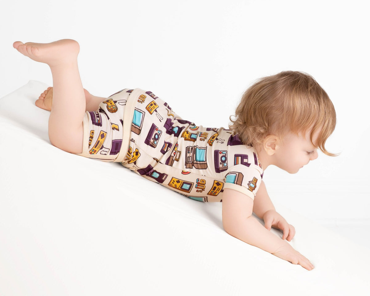 A toddler dressed in the Jammie Session 90's Retro Short Pajama Set is crawling down a white slope against a plain background.
