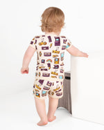 A toddler with light brown hair, dressed in the Jammie Session 90's Retro Short Pajama Set made of breathable bamboo, stands barefoot by a white piece of furniture, facing away from the camera.