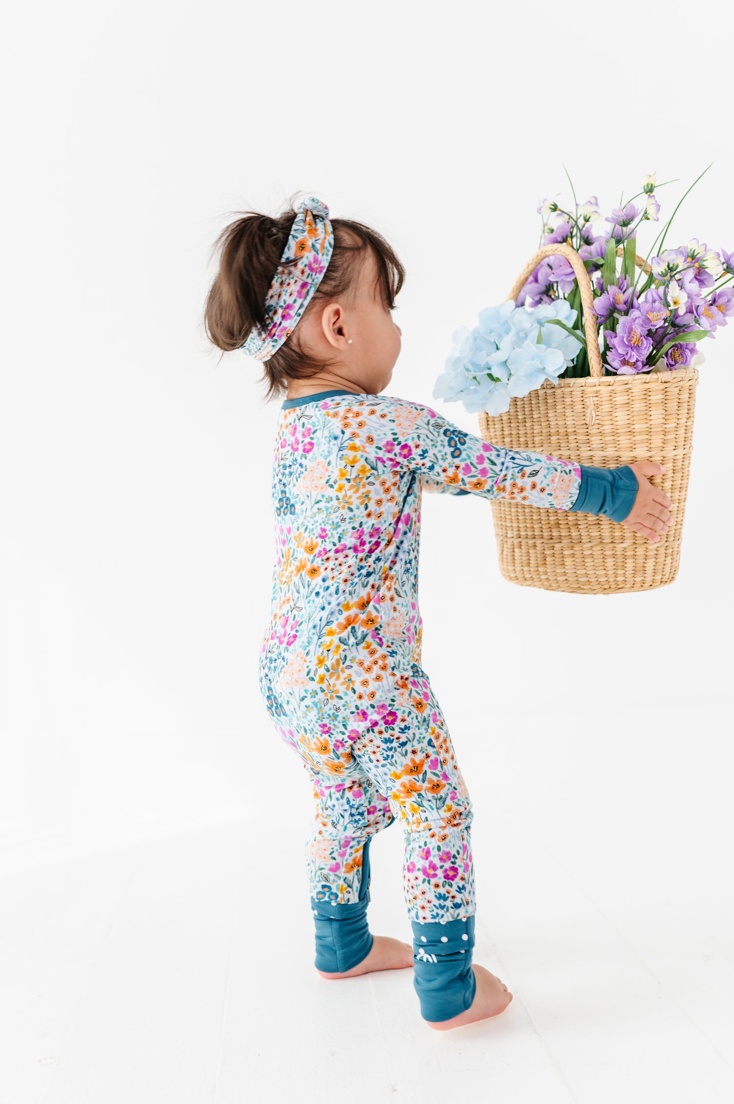 A toddler wears the Jammie Session Whispering Wildflowers Romper and a headband, holding a wicker basket of purple and blue flowers against a white background.