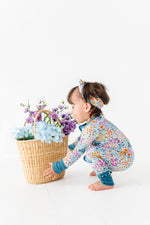 A toddler wears the Jammie Session Whispering Wildflowers Romper and headband, squatting with a wicker basket of purple and blue artificial flowers against a white background.
