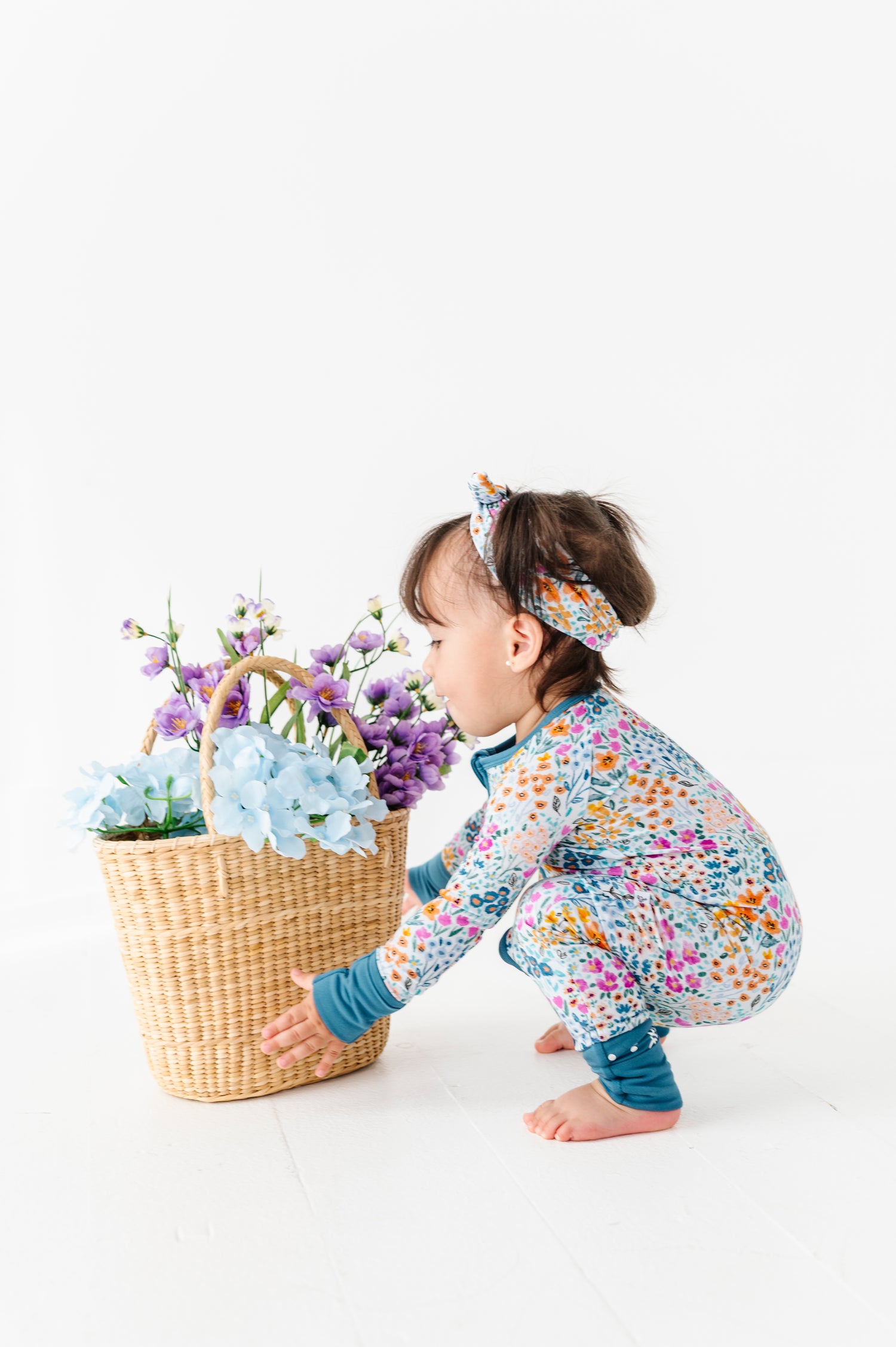 A toddler wears the Jammie Session Whispering Wildflowers Romper and headband, squatting with a wicker basket of purple and blue artificial flowers against a white background.
