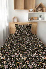 A neatly made bed features the Jammie Session Mystic Mushrooms Twin Fitted Sheet and Pillowcase Set, with a vibrant mushroom and foliage pattern; wooden shelves, decor, and a toy train are in the background.