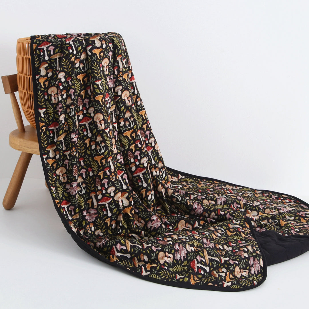 The Jammie Session Mystic Mushrooms Toddler Quilted Blanket, featuring a colorful mushroom and foliage pattern on black, is draped over a wooden chair against a white background.