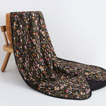 The Jammie Session Mystic Mushrooms Adult Quilted Blanket, featuring a vibrant mushroom and foliage design, is draped over a wooden chair against a plain white background.