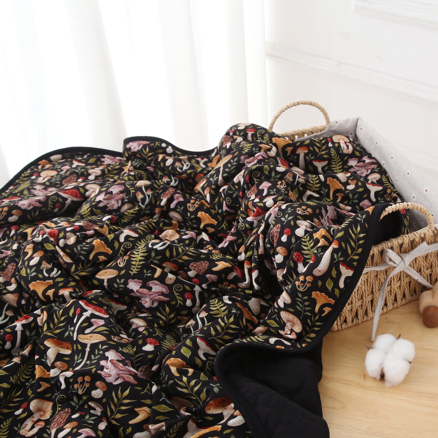 A woven basket with a gray cushion holds the Jammie Session Mystic Mushrooms Adult Quilted Blanket, a black hypoallergenic throw adorned with colorful mushroom and foliage designs, partially draped over the edge.