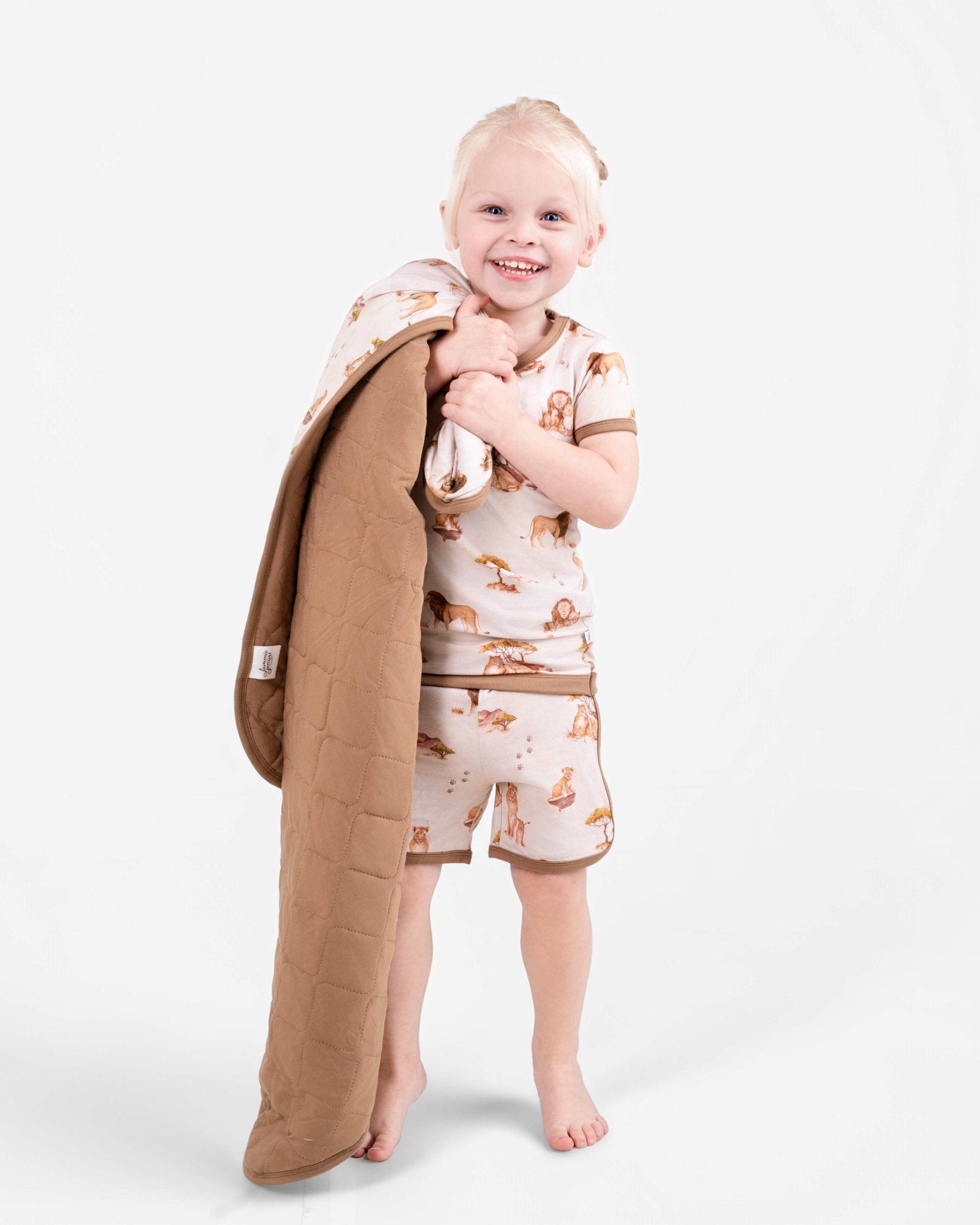 A smiling young child in the Ari (Lion) TENCEL™ Modal Short Pajama Set by Jammie Session holds a tan quilted blanket made from buttery soft, hypoallergenic fabric against a plain white background.