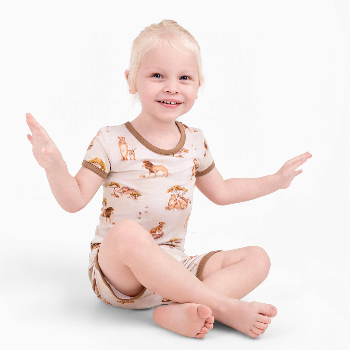 A young blond child sits cross-legged, smiling with hands up, wearing Jammie Session's Ari (Lion) TENCEL™ Modal Short Pajama Set—hypoallergenic, buttery soft pajamas featuring animal prints and brown trim.