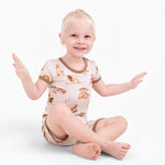A young blond child sits cross-legged, smiling with hands up, wearing Jammie Session's Ari (Lion) TENCEL™ Modal Short Pajama Set—hypoallergenic, buttery soft pajamas featuring animal prints and brown trim.