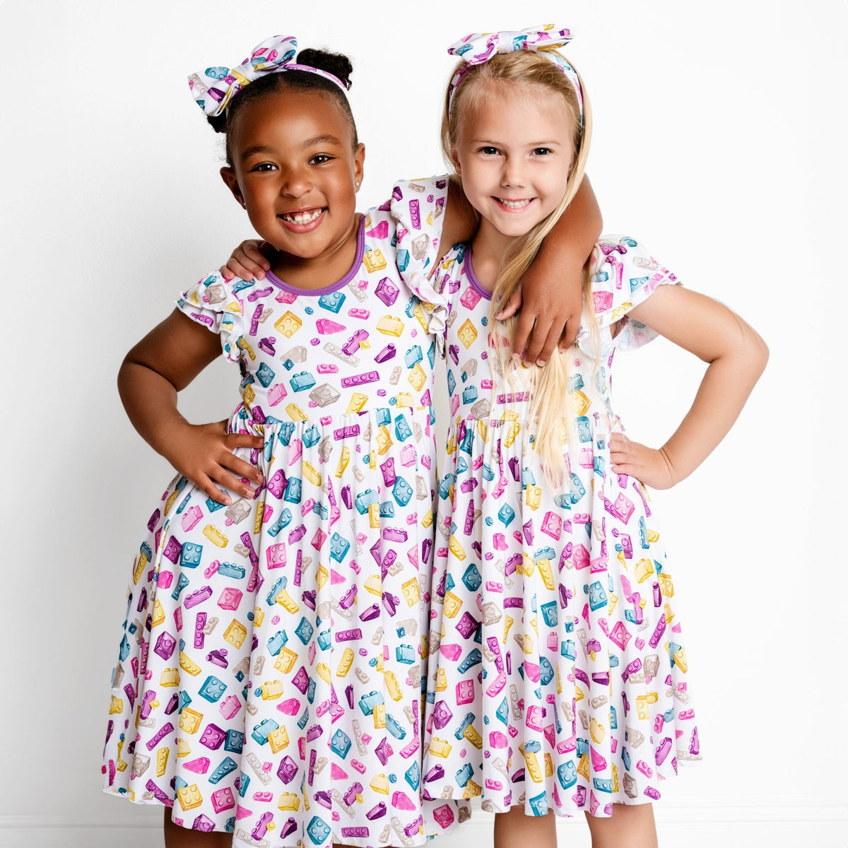 Two young girls smile side by side against a white background, wearing matching Block Party (Purple) Short Sleeve Big Kid Twirl Dresses by Big Dreams Little Jammies—soft, hypoallergenic bamboo outfits perfect for sensitive skin.