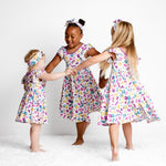 Three young girls wearing Big Dreams Little Jammies' Block Party (Purple) Short Sleeve Big Kid Twirl Dress hold hands and dance in a circle on a white, fluffy rug against a plain white background.