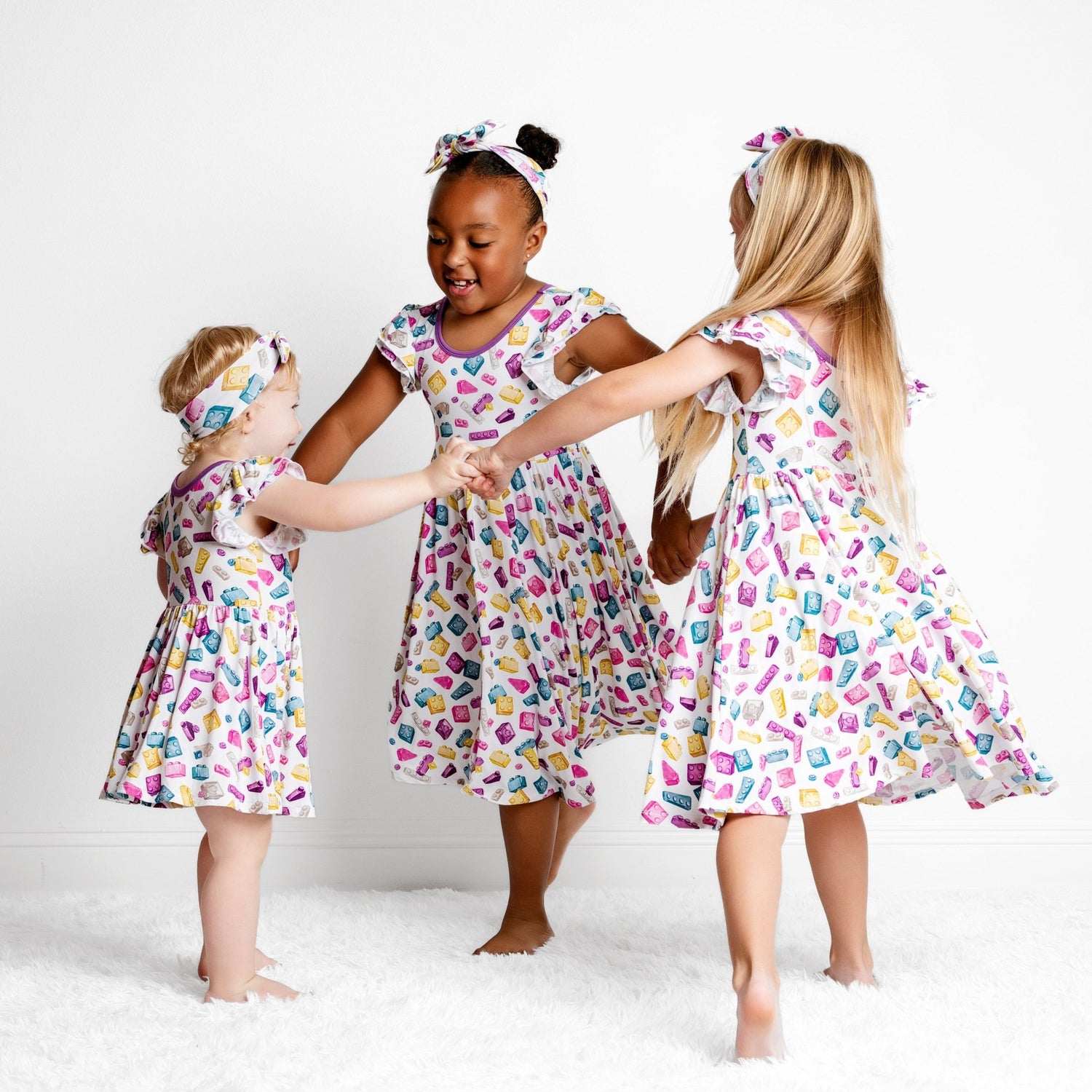 Three young girls wearing Big Dreams Little Jammies' Block Party (Purple) Short Sleeve Big Kid Twirl Dress hold hands and dance in a circle on a white, fluffy rug against a plain white background.